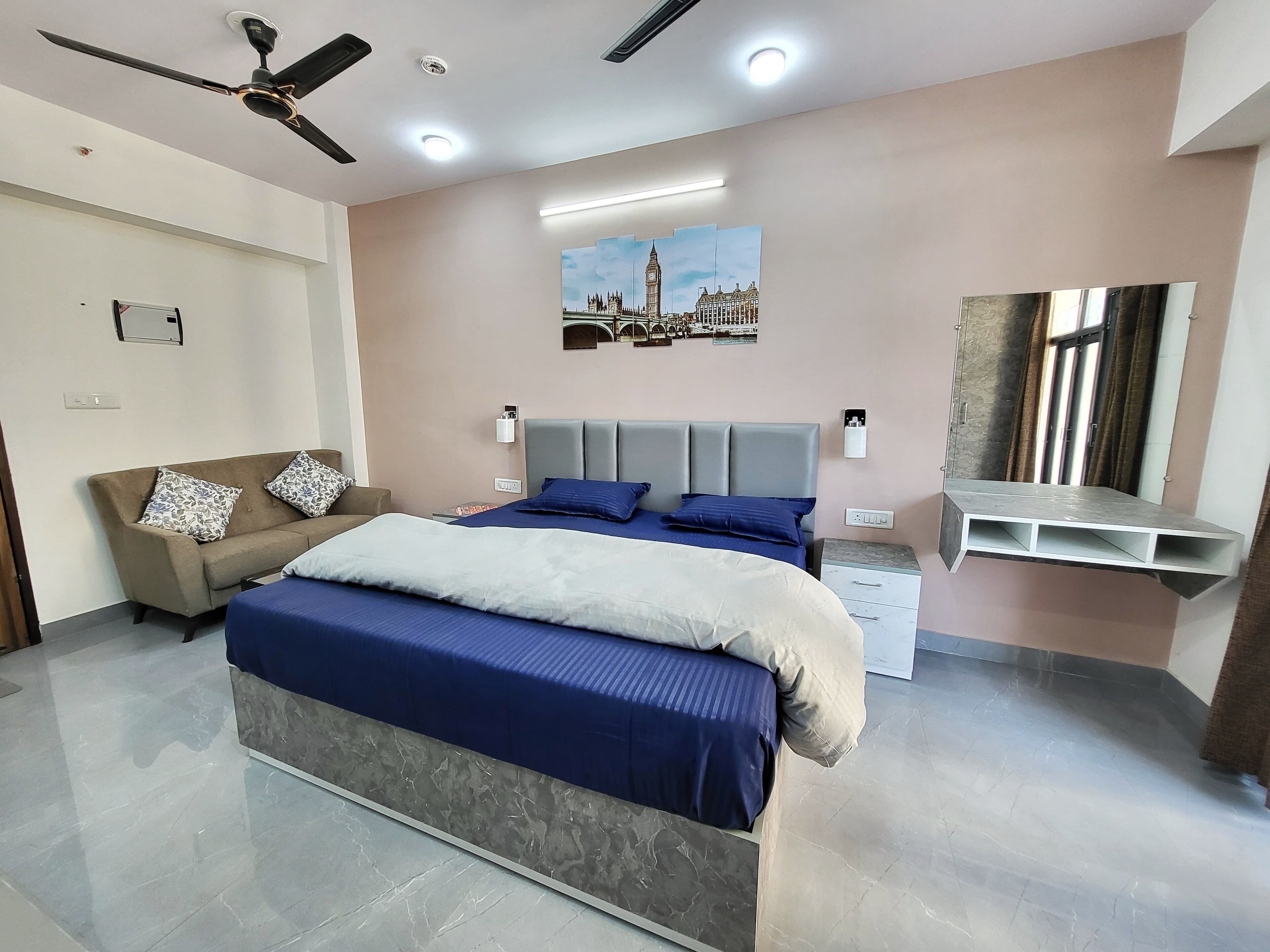 A bedroom with a bed, a sofa, a ceiling fan, a wall-mounted shelf, and a painting of a cityscape.