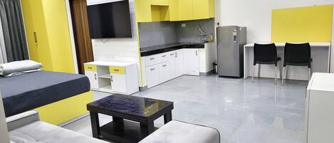 A modern hotel room with a sofa, a small table, a bed, a TV, a kitchen area with yellow cabinets, and a refrigerator.
