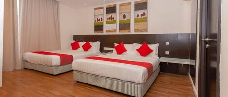 Quarto de hotel com duas camas, cada uma com almofadas vermelhas e roupa de cama listrada. Três quadros pendurados na parede acima das camas, e um aparelho de ar condicionado fixado na parede.