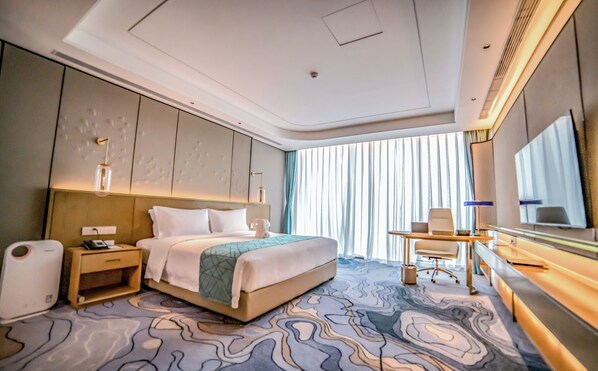 Una habitación de hotel moderna con una cama grande, un escritorio con una silla, una mesita de noche, un espejo amplio y vistas de la ciudad a través de ventanas de gran altura.