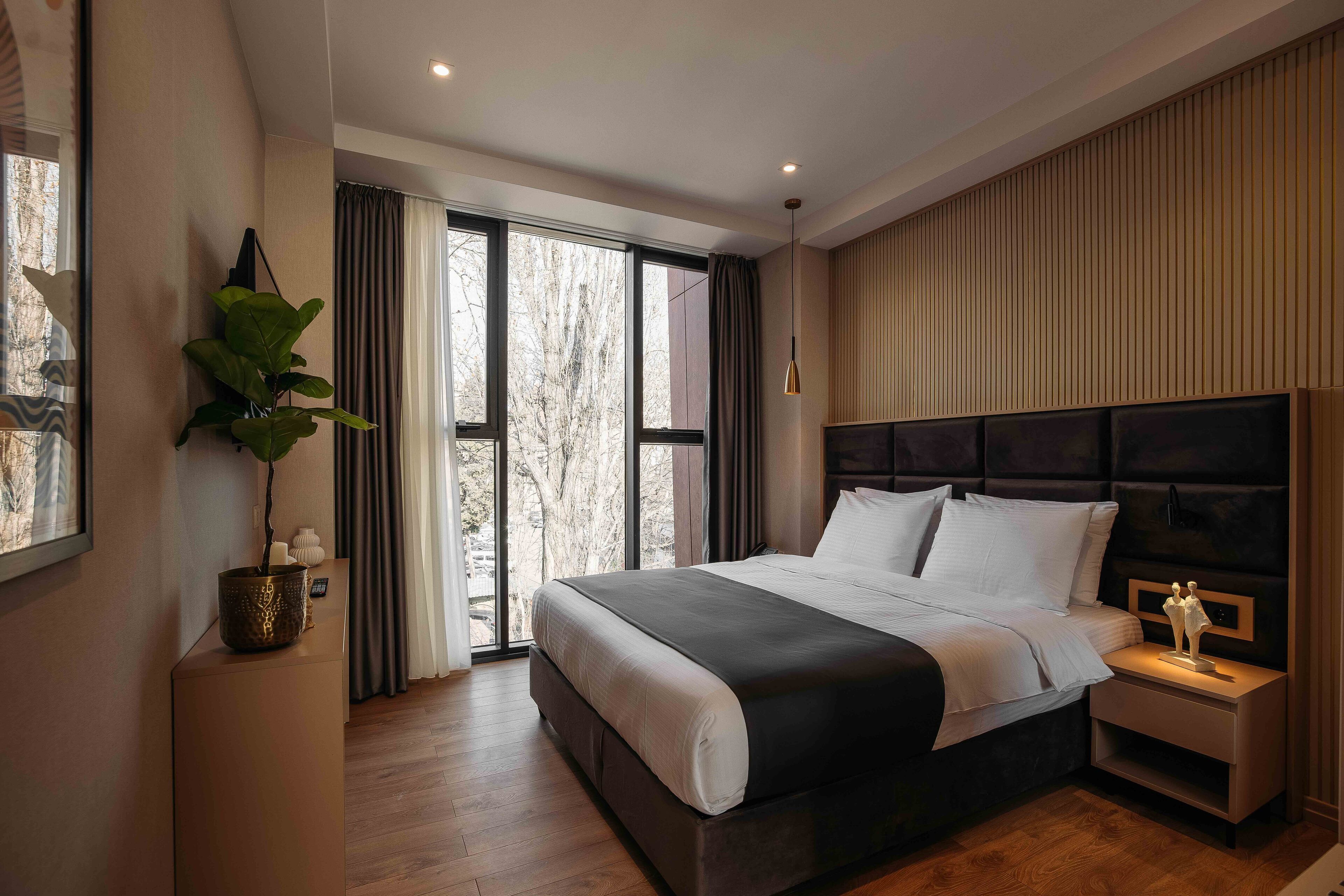 Une chambre moderne avec un grand lit, une table de chevet, une plante en pot et une vue sur les arbres par la fenêtre.