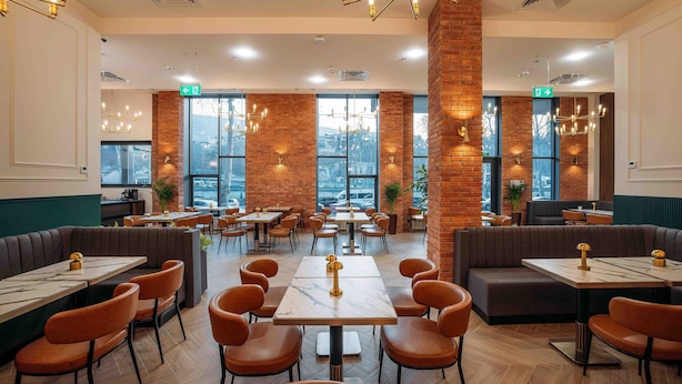 Un restaurant spacieux avec de hauts plafonds, des murs en briques apparentes et de grandes fenêtres offrant une vue sur la ville. Les tables et les banquettes sont disposées pour accueillir les clients.