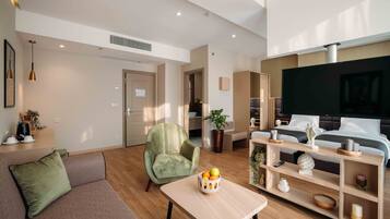 A modern living room with a sofa, armchair, and a coffee table with fruit. There is a TV and a bed in the background.