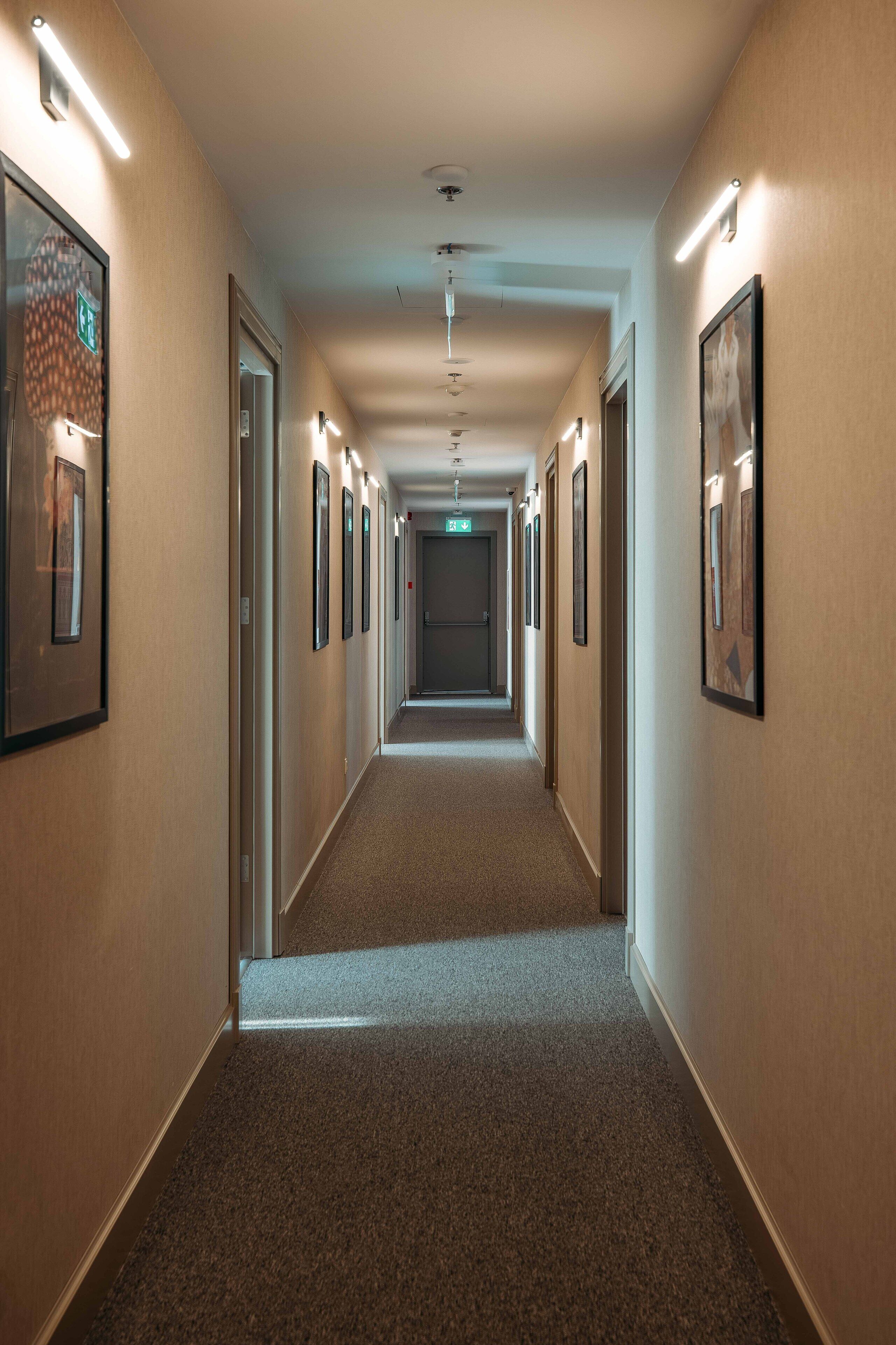 A long, narrow hotel corridor with framed pictures on the walls, carpeted floors, and a ceiling light fixture.