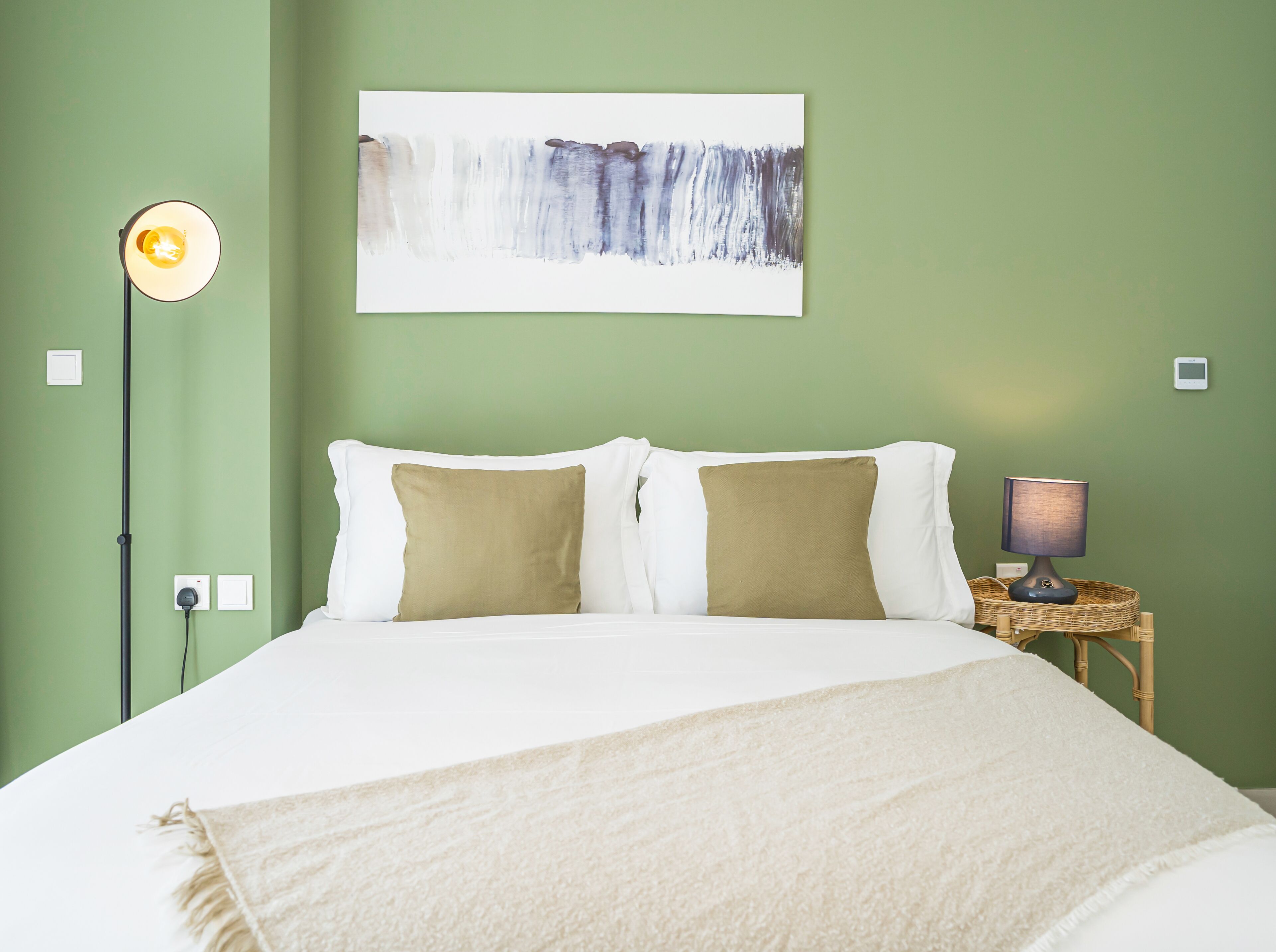 Een bed met witte en beige kussens, een witte deken, een vloerlamp en een abstract schilderij aan de muur.