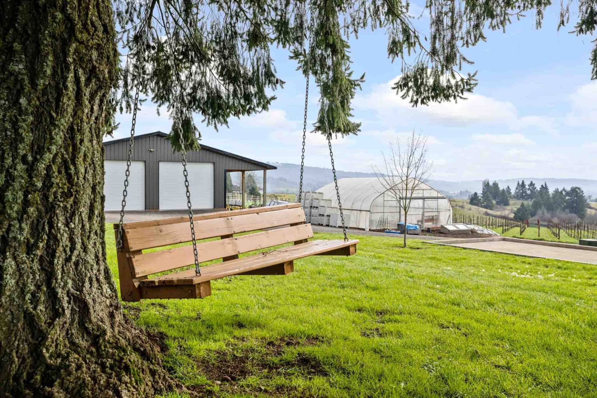 A wooden bench under a tree, a building with a garage, and a greenhouse in a grassy field.