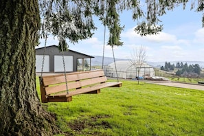 A wooden bench under a tree, a building with a garage, and a greenhouse in a grassy field.