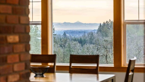 A dining area with a table and chairs, a brick wall, and a view of a forested landscape through large windows. - Picturesque Vineyard and Valley Views, Quiet and Private Setting, Huge Deck, *Complimentary Wine* (Newberg)