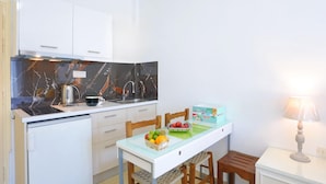 Una cocina compacta con armarios blancos, un protector contra salpicaduras de mármol, una pequeña mesa de comedor con sillas, un cuenco de frutas y una lámpara en una mesita de noche.
