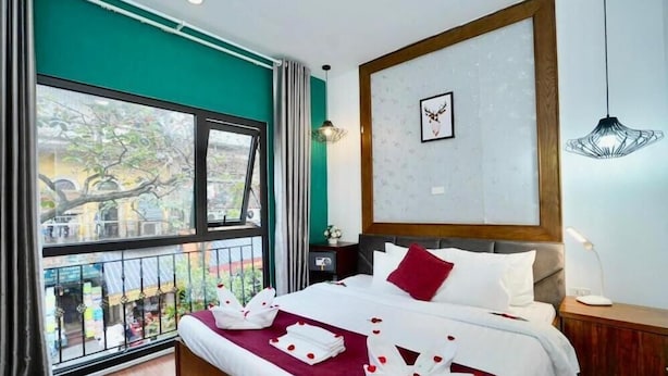 Una habitación de hotel con un ventanal grande, una cama con sábanas blancas y almohadas rojas, un cabecero de madera y una mesita de noche con una lámpara.