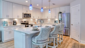Una cocina moderna con armarios blancos, electrodomésticos de acero inoxidable, una isla central con taburetes y luces colgantes.