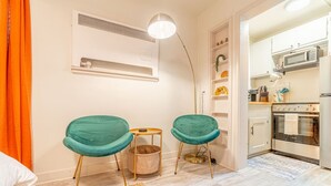 A compact living space with two teal chairs, a small side table, a woven rug, a white air conditioning unit, and a kitchen area with modern appliances. - Riverside Luxe: Stylish Designer Retreat (Reno)