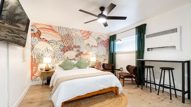 Une chambre à coucher comprenant un lit, un ventilateur de plafond, un bureau avec deux tabourets, une chaise et un climatiseur fixé au mur.