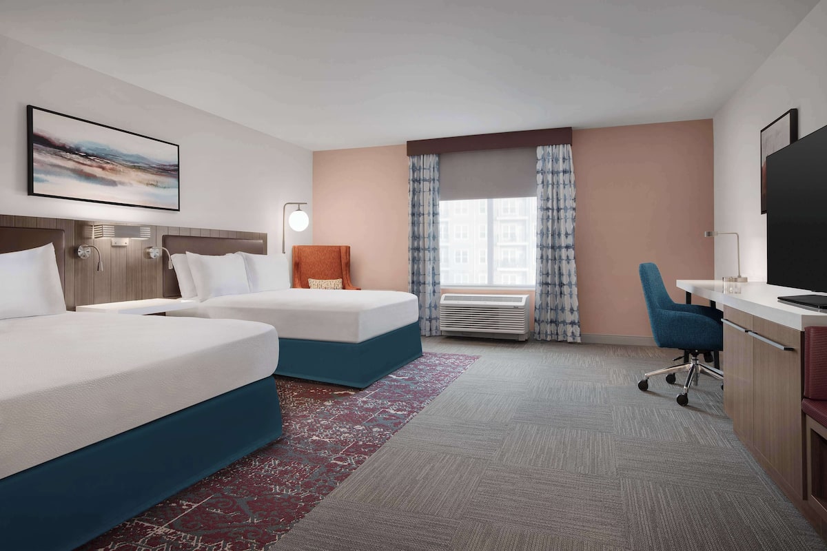 A hotel room with two beds, a desk, a chair, a TV, and a window with curtains.