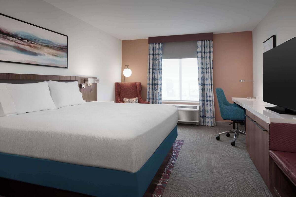 A hotel room with a large bed, a desk with a computer, a chair, a TV, and a window with curtains.