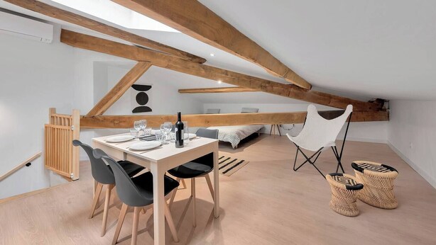 A modern loft bedroom with exposed wooden beams, a dining area with a table and chairs, and a cozy seating area with a chair and baskets. - Le Blackwood (Mazamet)
