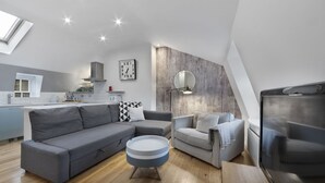 Un salon moderne avec un canapé gris, une table basse ronde et un fauteuil à carreaux. On aperçoit une cuisine à l’arrière-plan, avec un plan de travail blanc et une cuisinière.