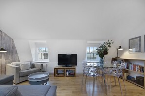 Ein modernes Loft-Apartment mit hohen Decken, einem Flachbildfernseher, einem Essbereich mit klaren Stühlen, einer Couch und einem Bücherregal.
