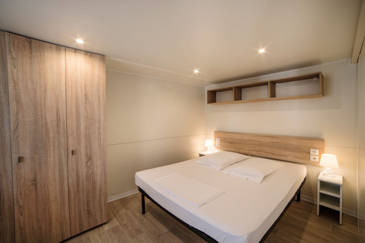 A bedroom with a large wardrobe, a bed with white linens, a nightstand with a lamp, and built-in shelving above the bed.