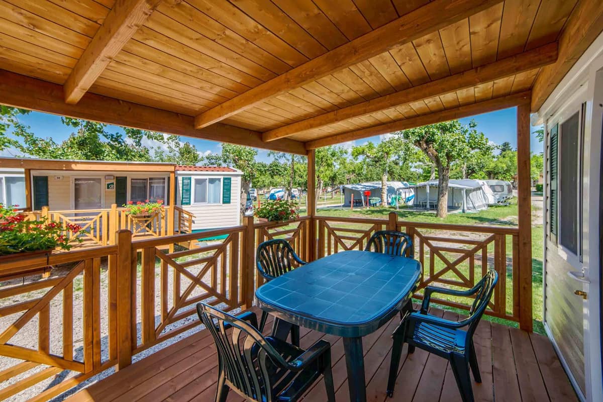 Una terraza cubierta al aire libre con una mesa y sillas, rodeada de barandillas de madera y con vista a casas móviles al fondo.