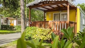 A yellow mobile home with a wooden porch and a covered area, surrounded by greenery and trees. - Baia Holiday Gasparina (Castelnuovo del Garda)