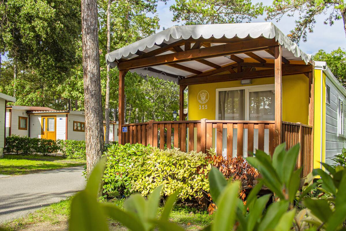Una casa móvil amarilla con porche de madera y un espacio cubierto, rodeada de follaje y árboles.