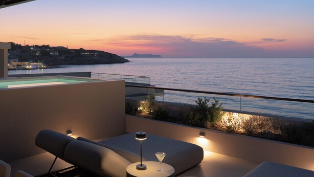 A balcony with a view of the sea at sunset, a lounge area with a glass table, and a pool.