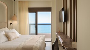 A hotel room with a bed, a TV mounted on the wall, a balcony with a view of the sea, and a wooden side table.