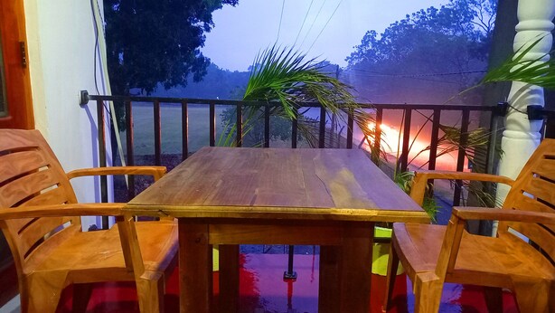 A wooden table with four chairs on a balcony with a railing, overlooking a street with trees and a sunset. - Sewana Guest INN (Wellawaya)