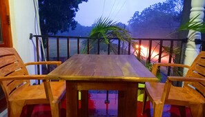 Une table en bois, entourée de quatre chaises, se trouve sur un balcon doté d’une balustrade, d’où l’on peut admirer une rue bordée d’arbres et un coucher de soleil.