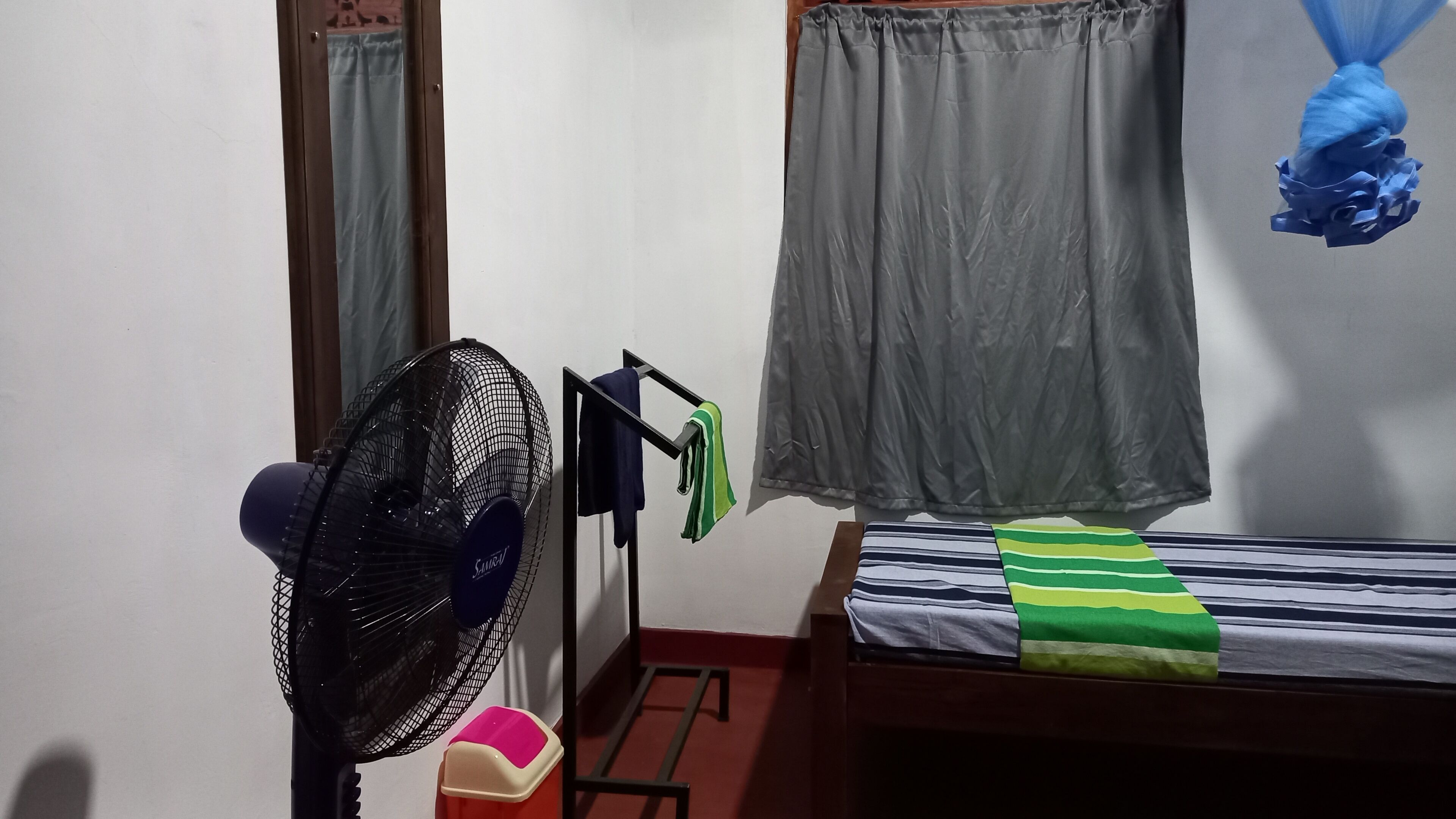 Et lille, enkelt møbleret værelse med en enkelt seng, en standventilator, et lille bord og et vindue med gardin.