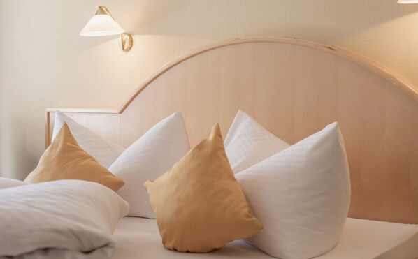 Uma cama bem arrumada com almofadas brancas e bege, uma cabeceira de madeira e uma luminária fixada na parede.