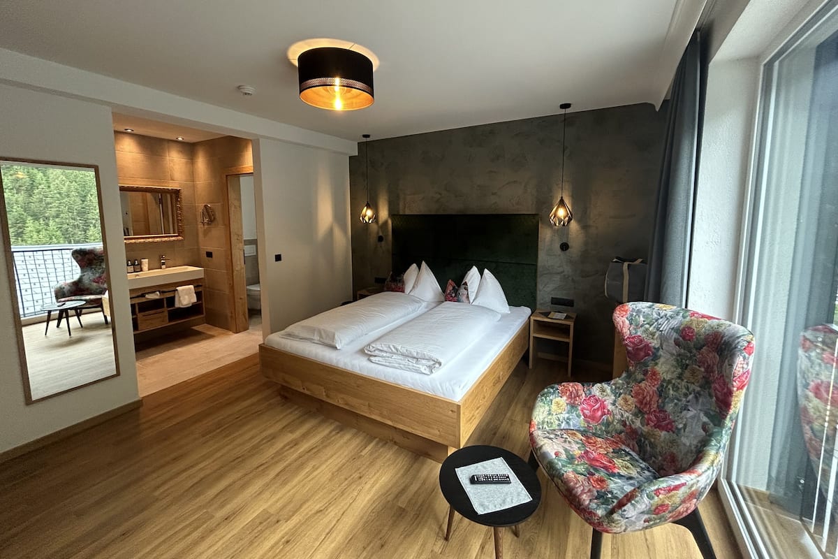 A modern hotel room with a wooden floor, a large bed with white linens, a floral armchair, a small side table, and a large window with a view of greenery.