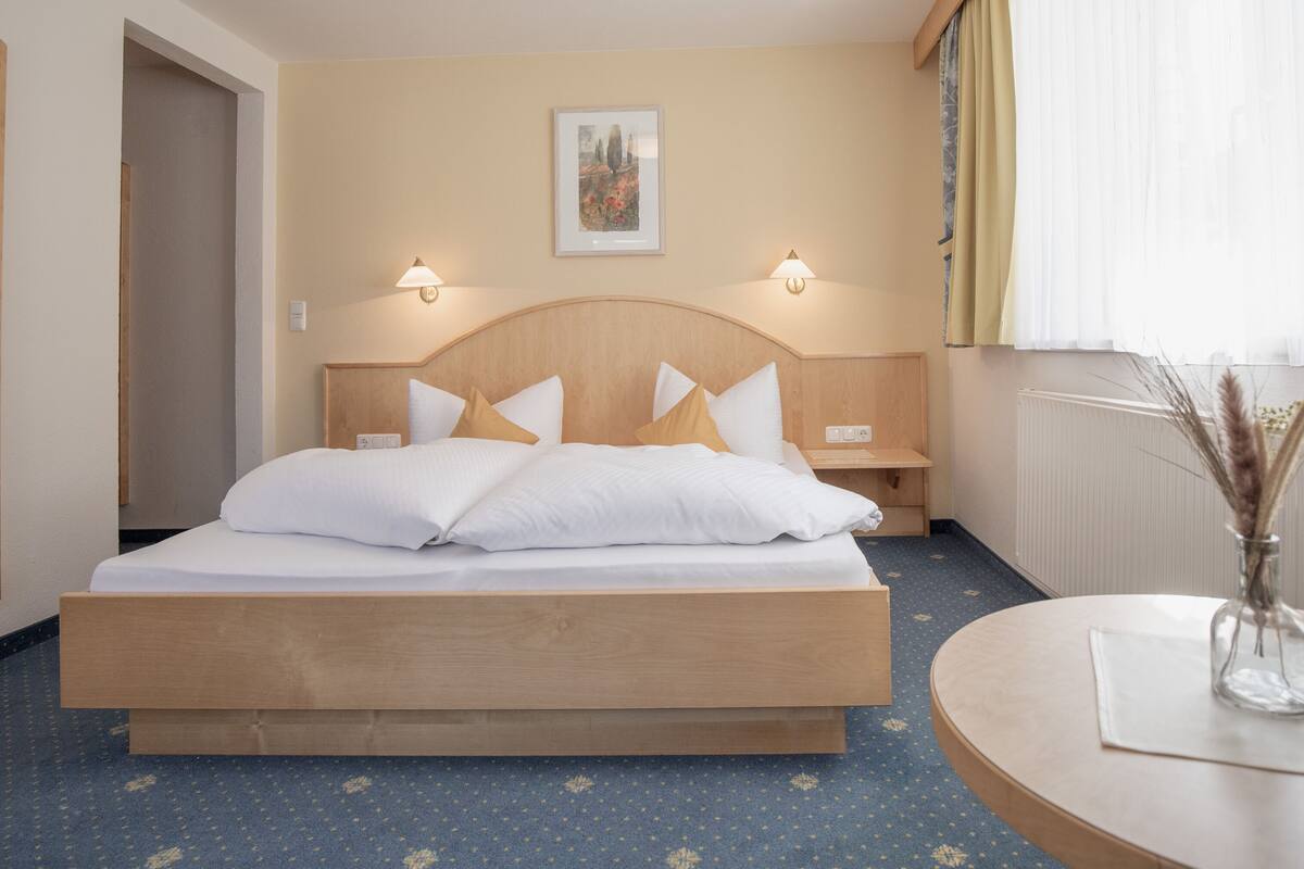 A hotel room with a large bed, bedside lamps, a round table with a vase, and a framed picture on the wall.