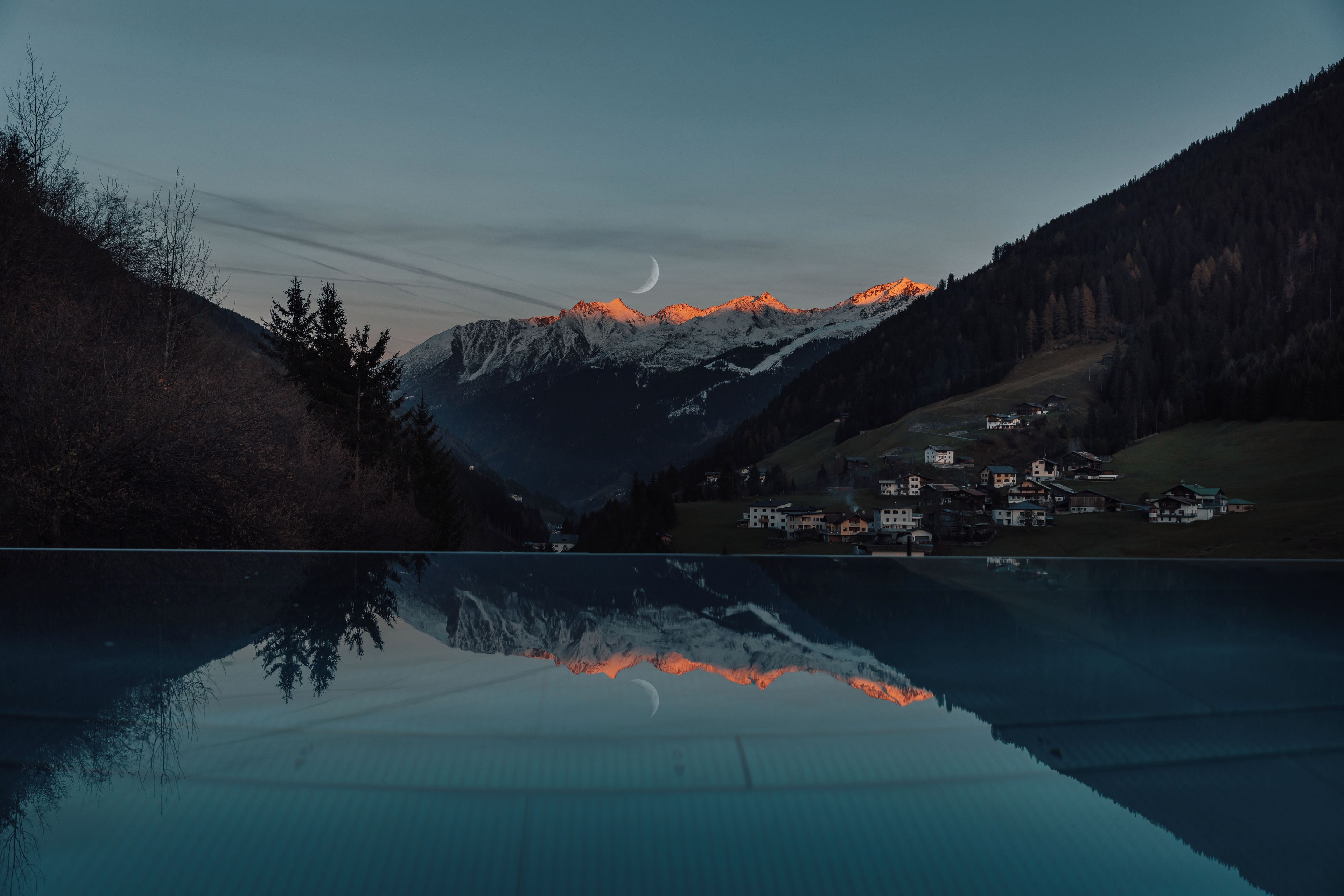 Berglandschaft mit einem See, der die Umgebung spiegelt, darunter schneebedeckte Gipfel und ein Halbmond.