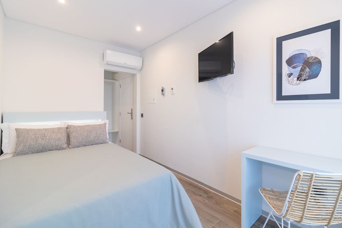A bedroom with a bed, a desk, a chair, a TV, and an air conditioning unit.