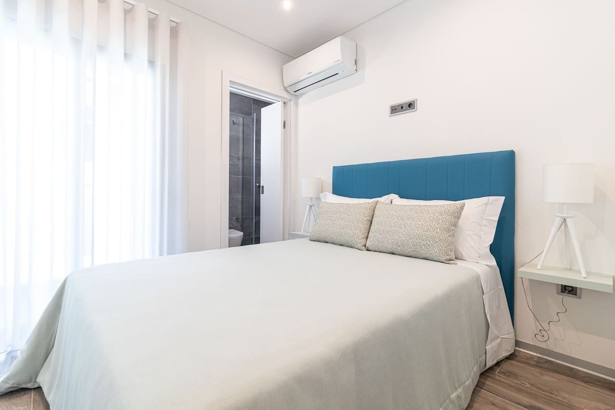 A modern hotel room with a large bed, a blue headboard, white bedding, a small bedside table with a lamp, and an air conditioning unit on the wall.
