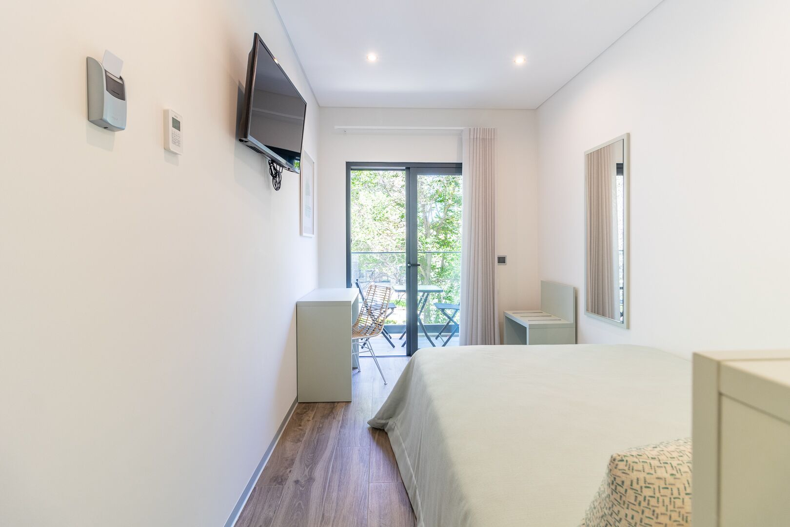 A hotel room with a bed, a TV mounted on the wall, a small table, and a chair. There is a balcony with a table and chairs outside the room.