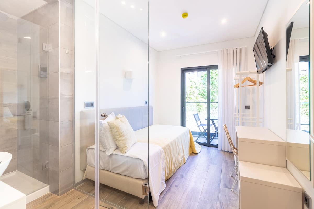 A modern hotel room with a glass shower enclosure, a bed with white bedding, a small table with a chair, and a TV mounted on the wall.