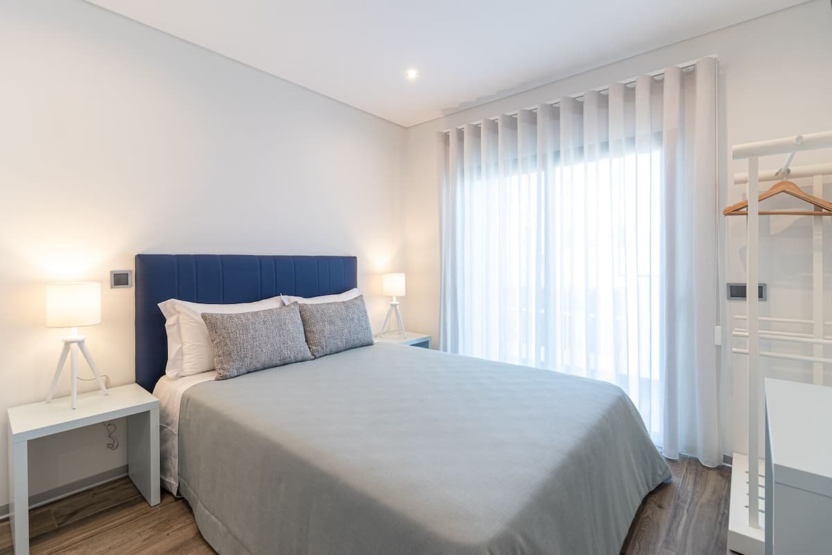 A modern hotel room with a large bed, two bedside tables with lamps, a wardrobe, and a large window with sheer curtains.
