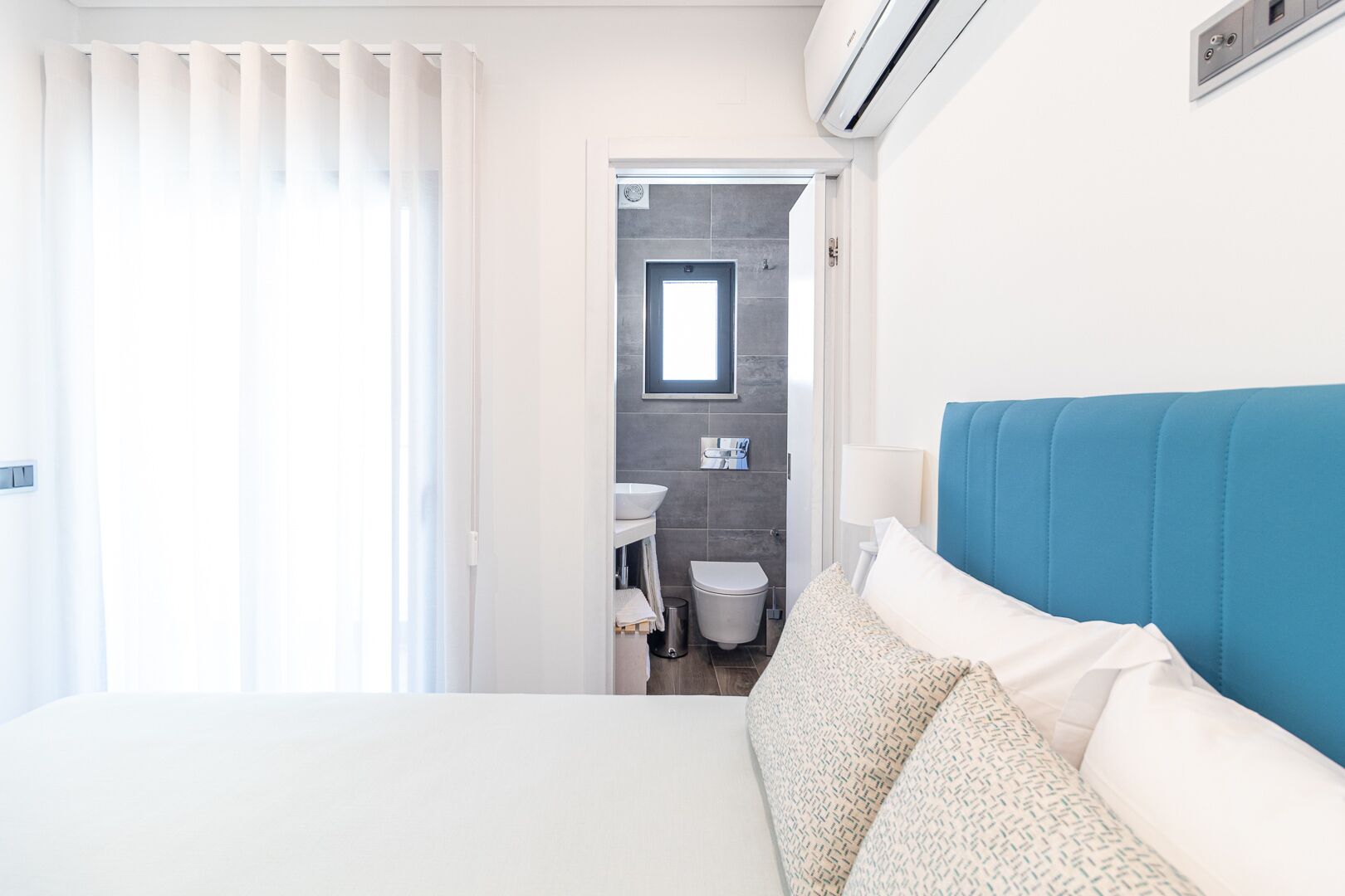 A hotel room with a bed, a bathroom visible through an open door, and an air conditioning unit on the wall.