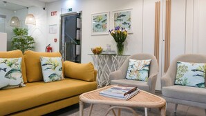 Zona de recepción con un sofá amarillo, sillones grises, una mesita con un libro, una mesa de cristal con un jarrón de flores y obras de arte enmarcadas en la pared.