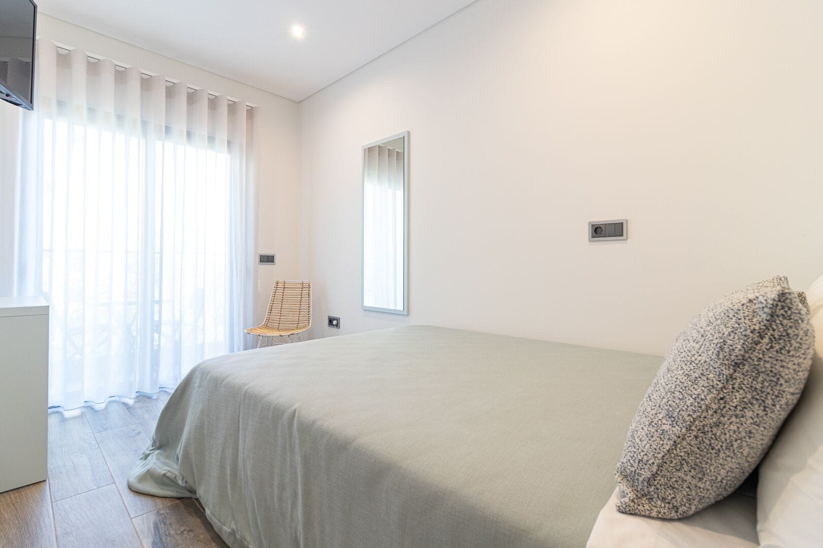 Quarto minimalista com uma cama grande, uma cadeira e uma janela com cortinas leves.