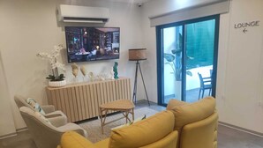 Una sala de estar moderna con un sofá amarillo, un sillón blanco y un televisor montado en la pared. Hay una mesita y una lámpara de pie. Se ve un cartel que indica un área de descanso en la pared.