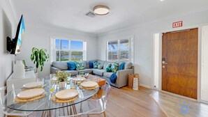 A modern living room with a glass dining table, a grey sectional sofa, and a wooden door marked "EXIT". - Beach Paradise : Family-Friendly + Free Parking (Hollywood)