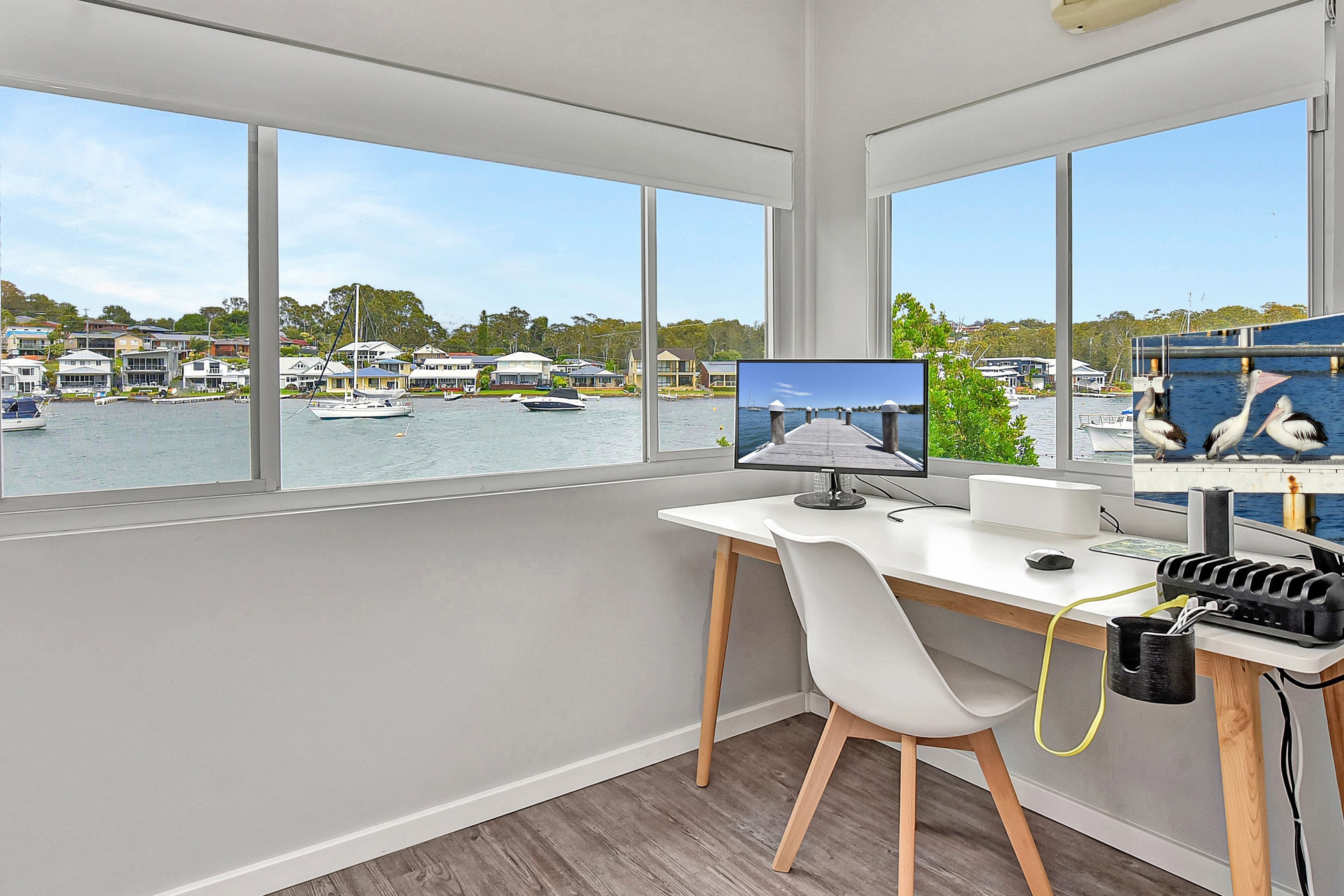 Ein moderner Arbeitsraum mit Schreibtisch, Stuhl und Computermonitor, mit Blick auf eine Marina mit Booten und Häusern.