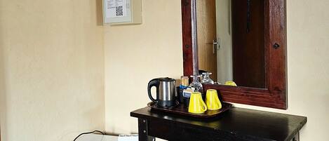 A compact room with a small white refrigerator, a wooden table with a mirror, and a tray with yellow cups and a kettle.