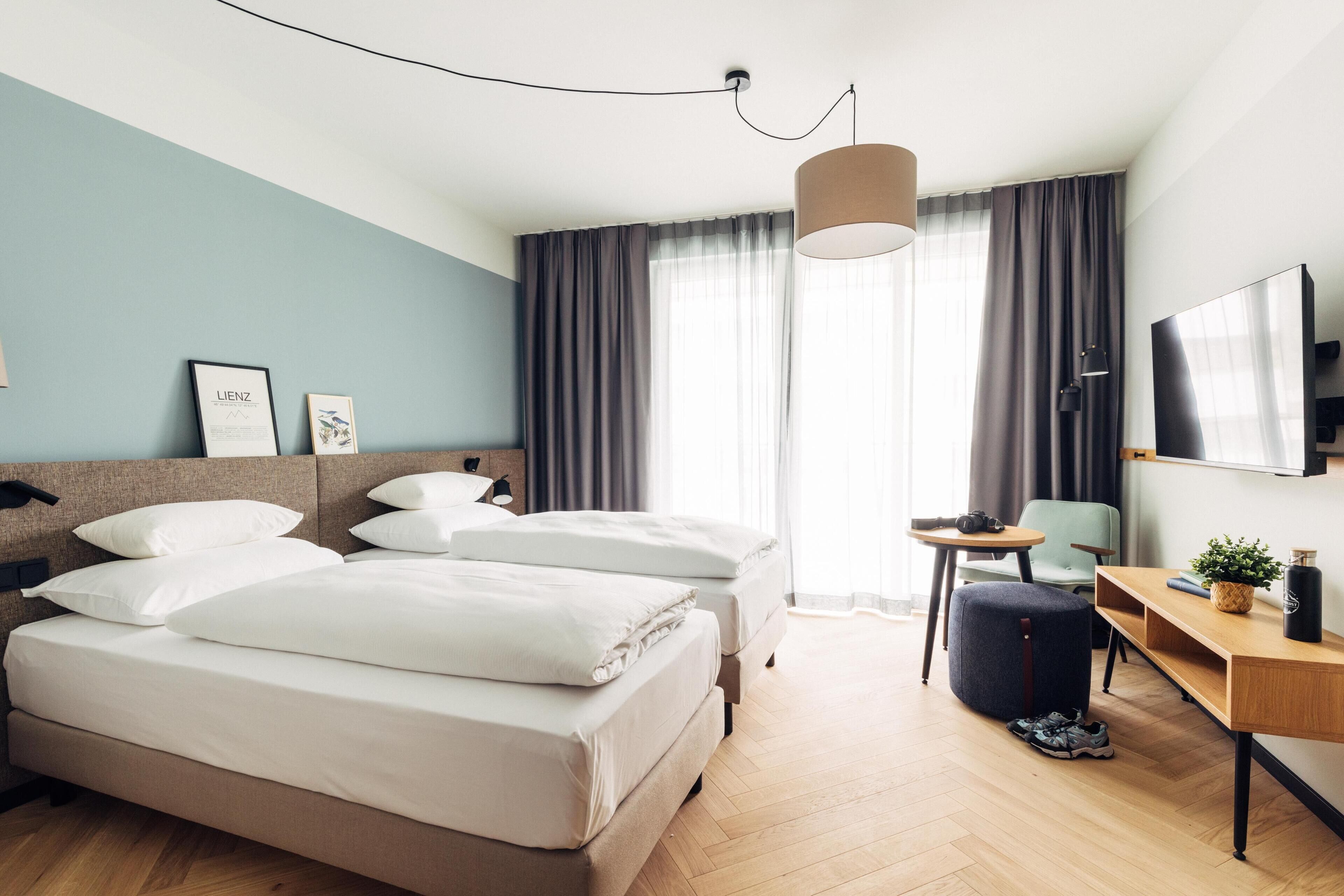 Une chambre d’hôtel moderne, équipée d’un lit, de tables de chevet, d’un bureau, d’une chaise et d’une grande fenêtre avec des rideaux.