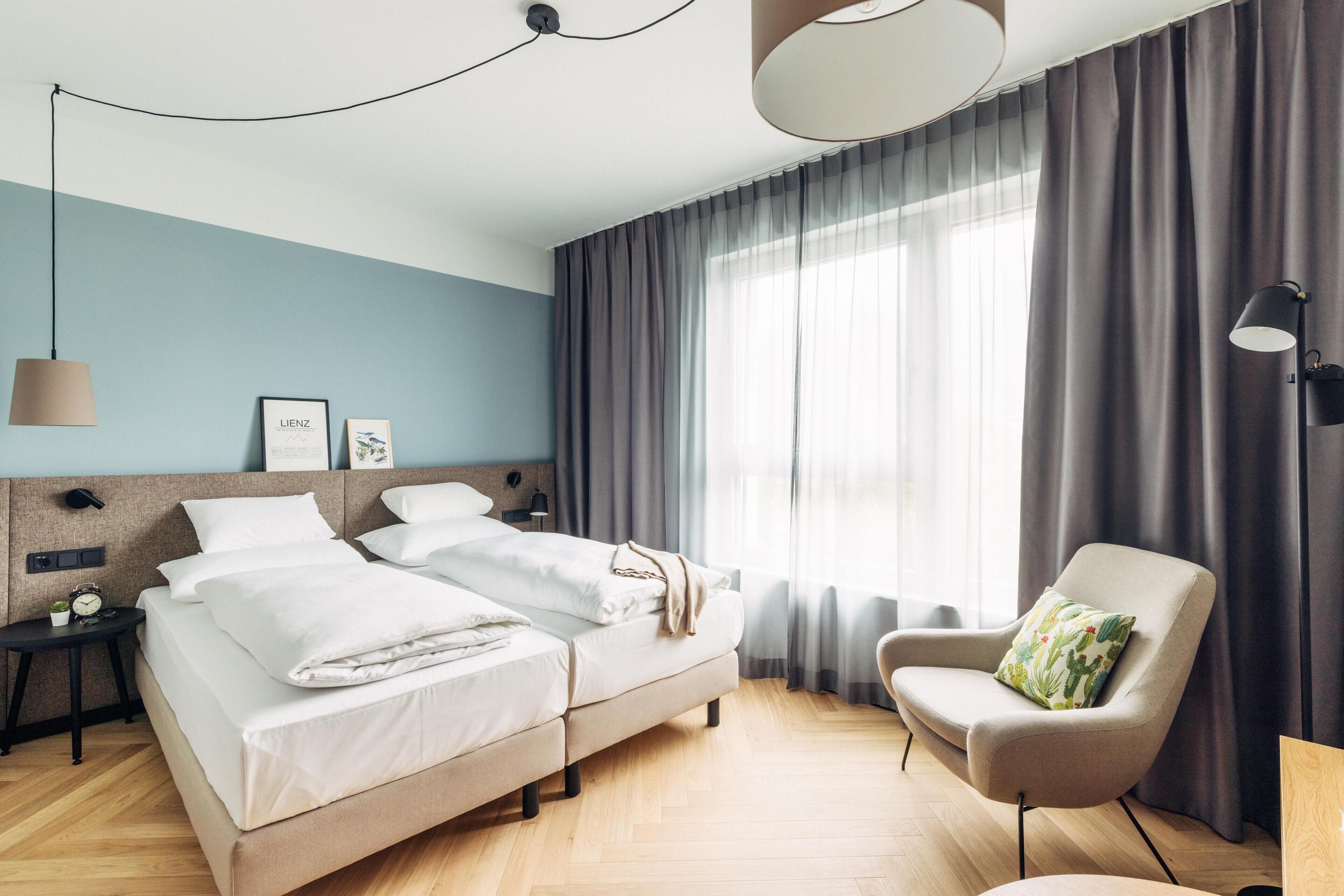 Ein modernes Hotelzimmer mit einem großen Bett, einem Sessel, einem Nachttisch und einem Fenster mit Vorhängen.