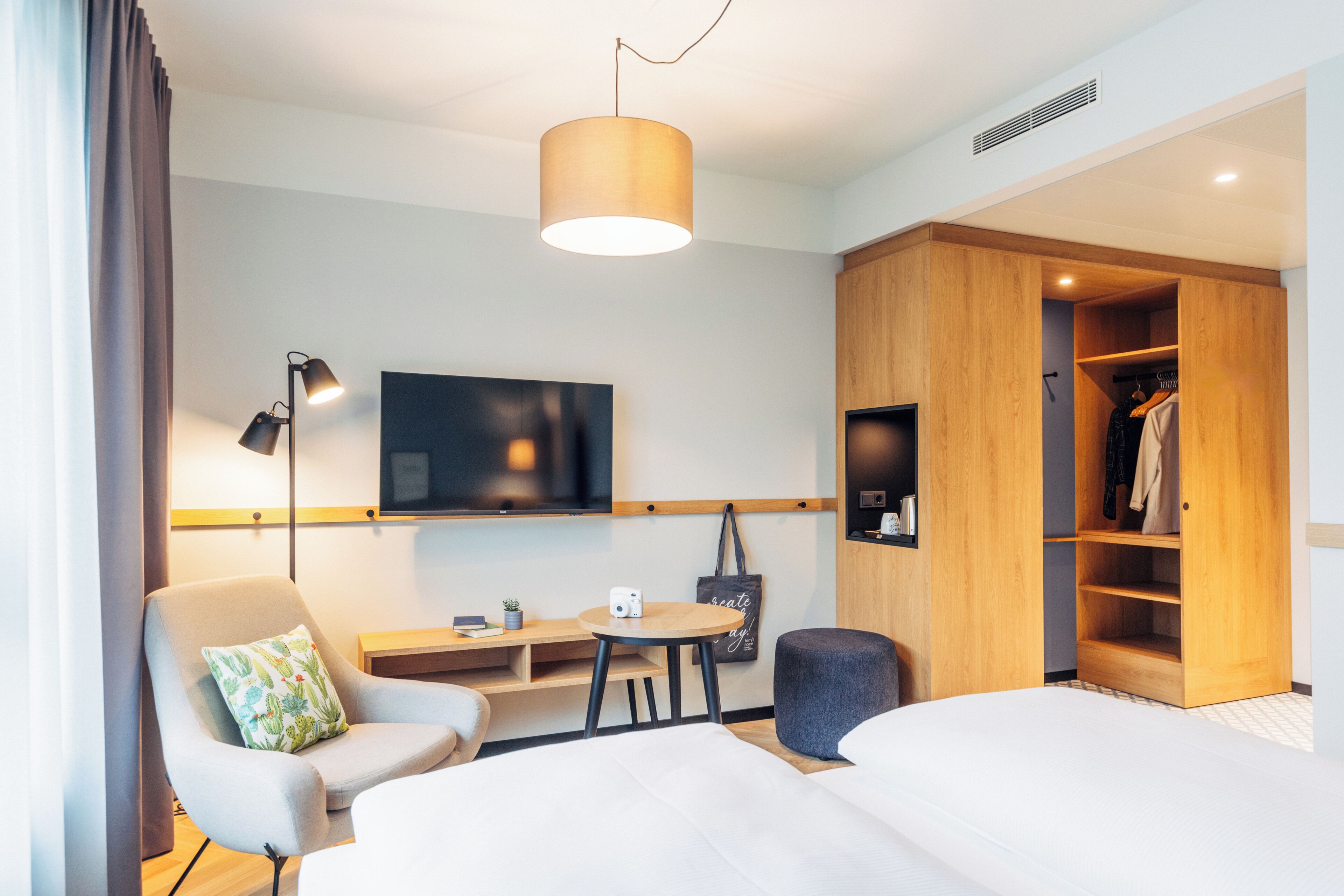 A modern hotel room with a bed, a desk, a chair, a TV, a wardrobe, and a hanging coat rack.
